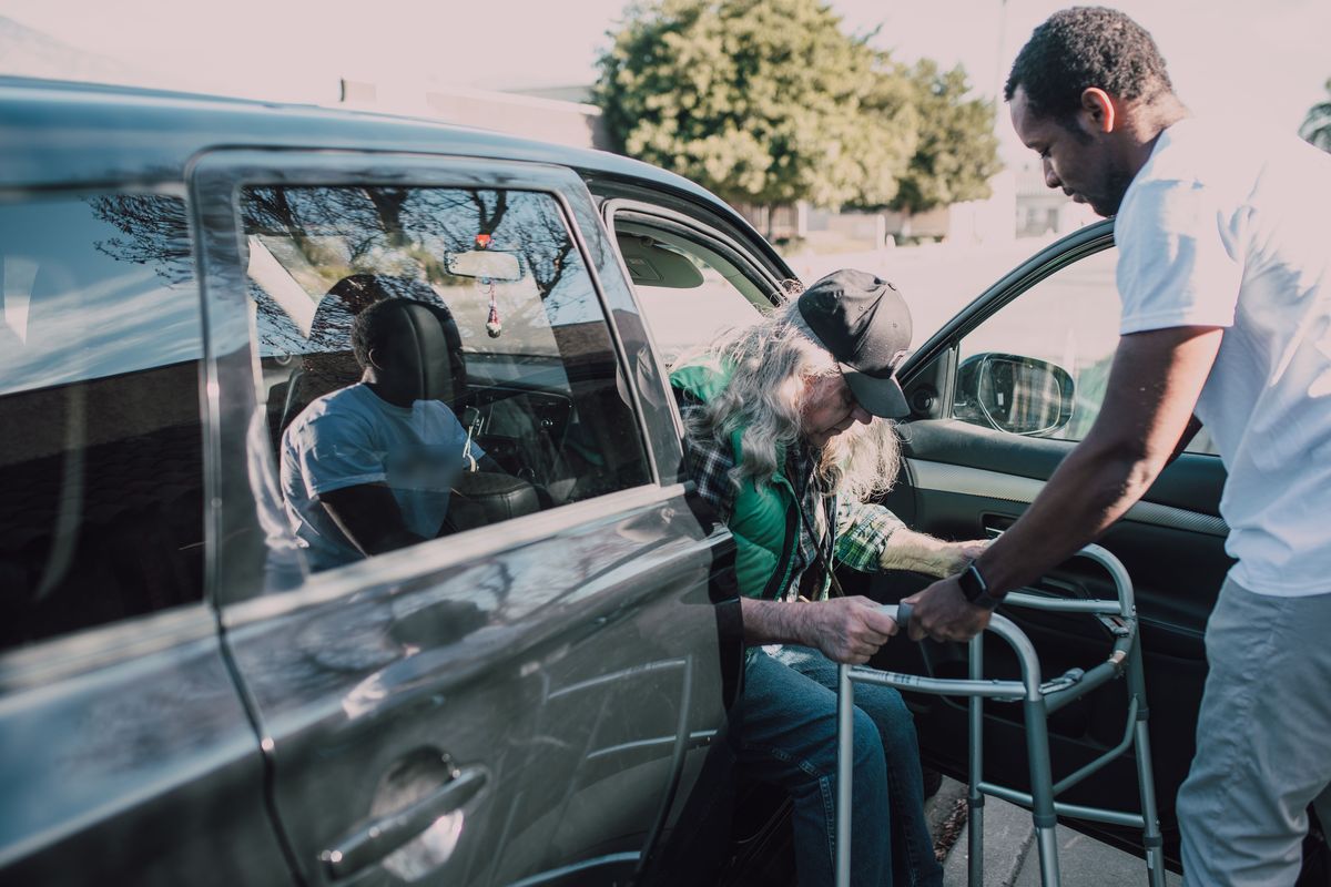 Een man helpt een oudere man met een rollator uit een wagen