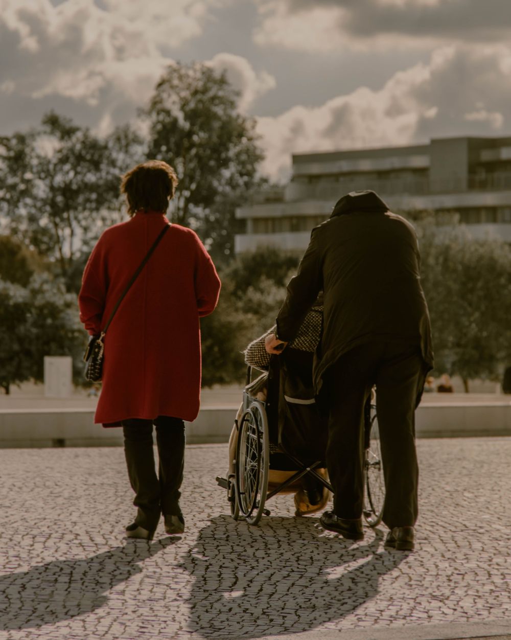 Twee mensen wandelen met iemand in een rolstoel over een plein, van achteren gezien