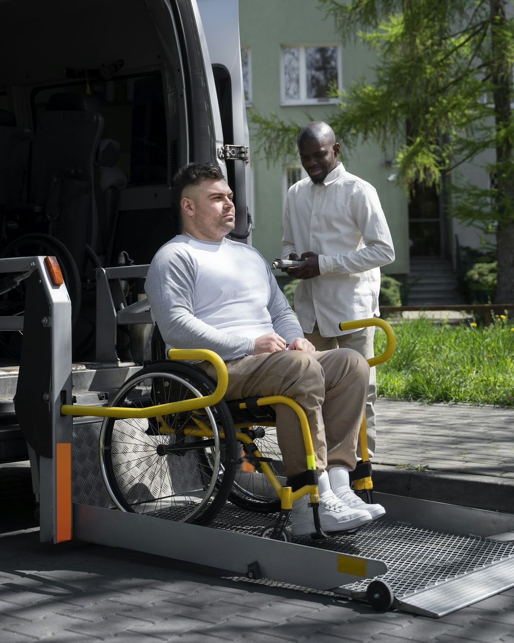 Een man in een rolstoel staat op de lift achteraan een bestelwagen, een begeleider bedient de lift
